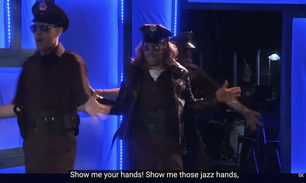 Sam Sweetly (Jeff Blim), a cop, does jazz hands while singing, “Show me your hands! Show me those jazz hands,” in the musical, “The Guy Who Didn’t Like Musicals.”