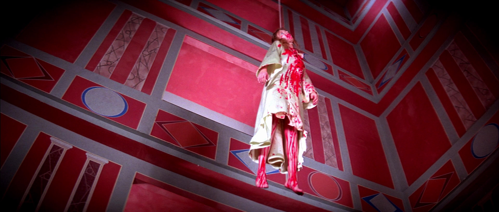 Pat (Eva Axen) is shown bloodied and hanging from a wire against a red background in Suspiria